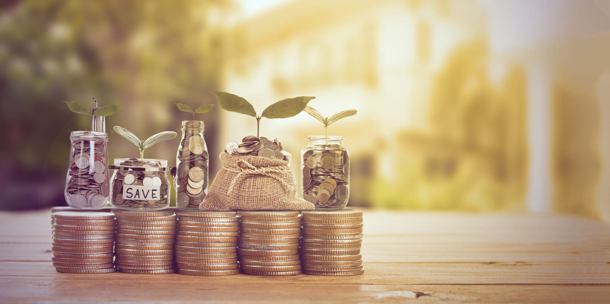 Three jars with coins and plants rise from stacked coins, symbolizing savings and investment growth on a wooden surface, with a soft, blurred background.
